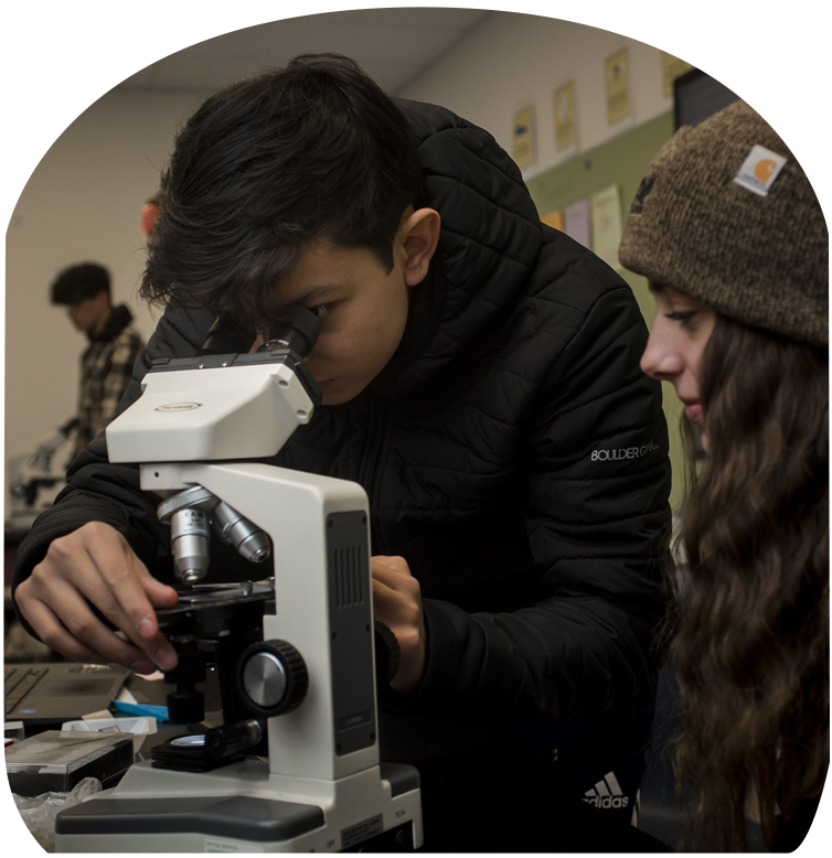 Two high school-aged students; one male and one female. The female student is observing the male student look through a white microscope.
